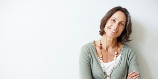 Reifere Frau mit halblangen braunen Haaren steht lächelnd mit verschränkten Armen vor einer weißen Wand, trägt eine lange Kette und einen hellgrünen Cardigan.
