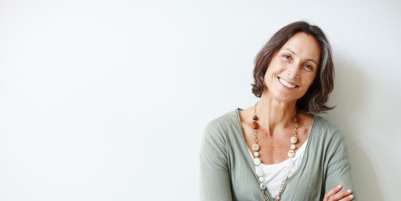 Reifere Frau mit halblangen braunen Haaren steht lächelnd mit verschränkten Armen vor einer weißen Wand, trägt eine lange Kette und einen hellgrünen Cardigan.