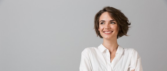 Junge Frau mit kurzem braunen Haar lächelt und verschränkt die Arme, vor grauem Hintergrund.