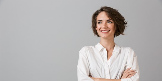 Junge Frau mit kurzem braunen Haar lächelt und verschränkt die Arme, vor grauem Hintergrund.