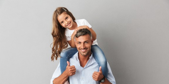 Glücklicher Vater mit seiner Tochter auf den Schultern, beide lächeln in die Kamera, vor grauem Hintergrund.
