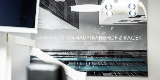 Wandbild mit moderner Architektur und dem Schriftzug „Zahnärzte am Hauptbahnhof // Racek – seit 2013“ in einer Zahnarztpraxis, im Vordergrund ein Teil der Behandlungseinheit.