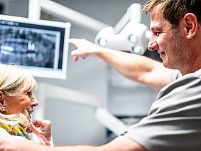 Zahnarzt zeigt einer älteren Patientin ein Röntgenbild auf einem Monitor und erklärt die Diagnose.